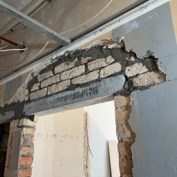 Daniella Deneka - Wzmocnienie nadproża nad otworem drzwiowym z widoczną konstrukcją z cegieł i zaprawy, częściowo obudowane płytą kartonowo-gipsową, z metalową konstrukcją sufitu podwieszanego w tle.