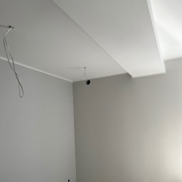 Daniella Deneka - Pomieszczenie po świeżym pomalowaniu ścian w odcieniach szarości, widoczne przewody elektryczne wystające z sufitu i małe otwory w ścianie na dole, surowa podłoga przygotowana do dalszych prac...