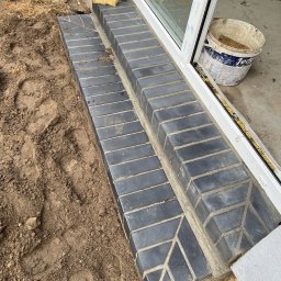 Daniella Deneka - Wykończone schody wejściowe z ciemnej cegły klinkierowej z dekoracyjnym wzorem ułożone przy białych drzwiach tarasowych, widoczne ślady ziemi i wiadro po zaprawie, Poznań.