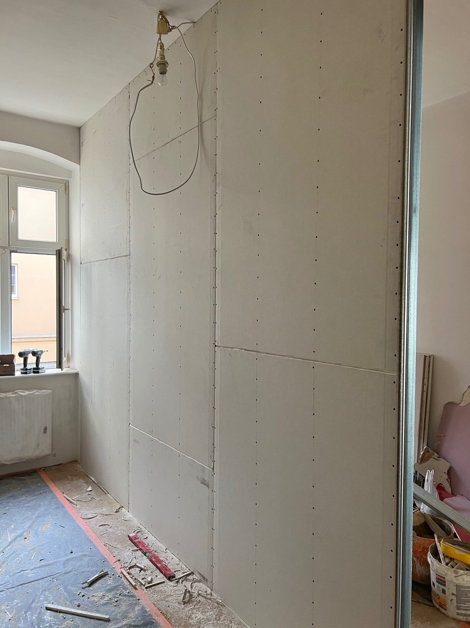 Ściana z płyt kartonowo-gipsowych przymocowana do metalowego stelażu, widoczne ślady po wkrętach, na podłodze folia ochronna, narzędzia przy ścianie, okno z grzejnikiem w tle.