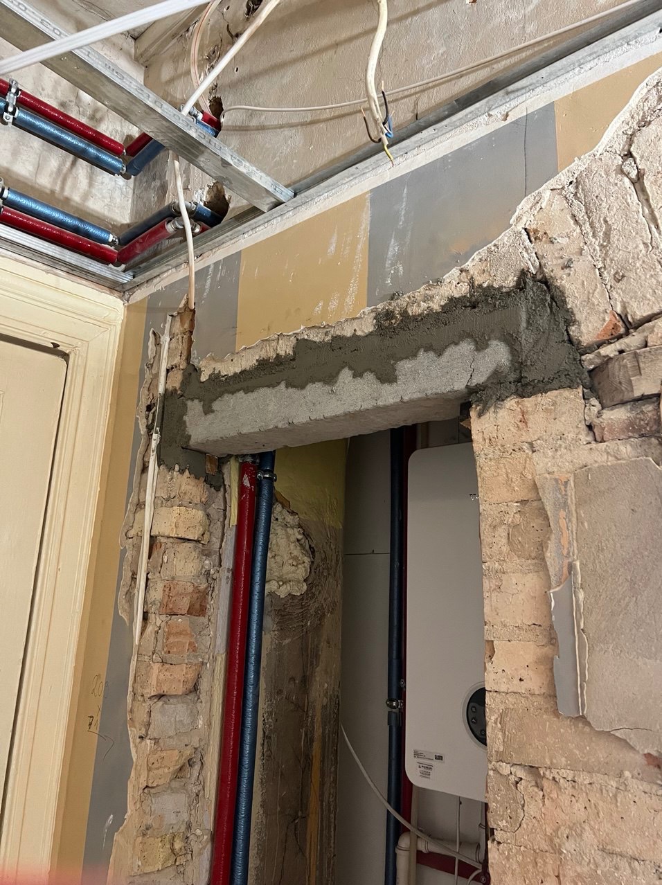 Ościeżnica drzwiowa w trakcie naprawy z widoczną zaprawą, odsłoniętą cegłą, rurami instalacyjnymi i fragmentem podwieszanego sufitu.