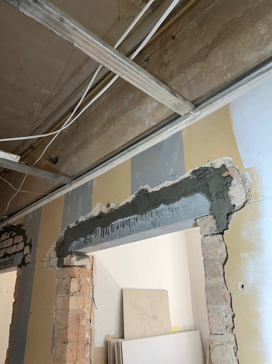 Prace remontowe: Przygotowanie otworu drzwiowego z nałożoną warstwą szpachli na ceglany mur, widoczne profile sufitowe i płyty karton-gips w tle.