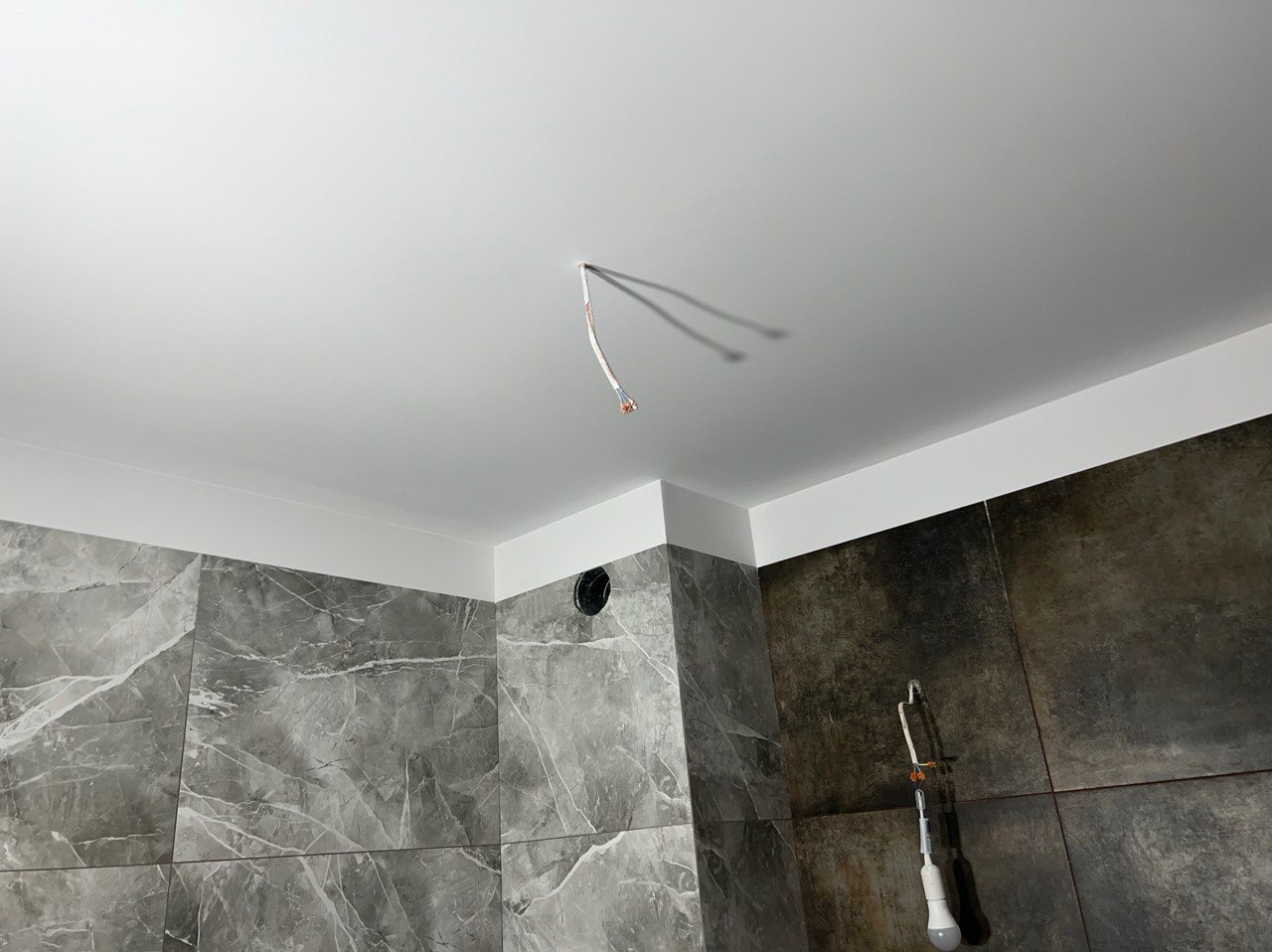 Niedokończone wnętrze łazienki z szarymi płytkami imitującymi marmur na ścianach, biały sufit z wystającymi kablami i otworem wentylacyjnym, oraz wiszącą lampą roboczą.