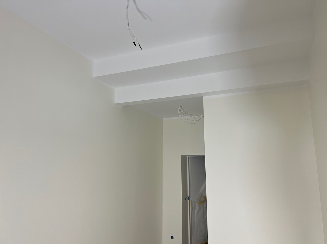 Świeżo pomalowane, jasne wnętrze z białymi ścianami i sufitem, widoczne elementy konstrukcyjne sufitu i wystające kable. W tle widoczne drzwi zabezpieczone folią.