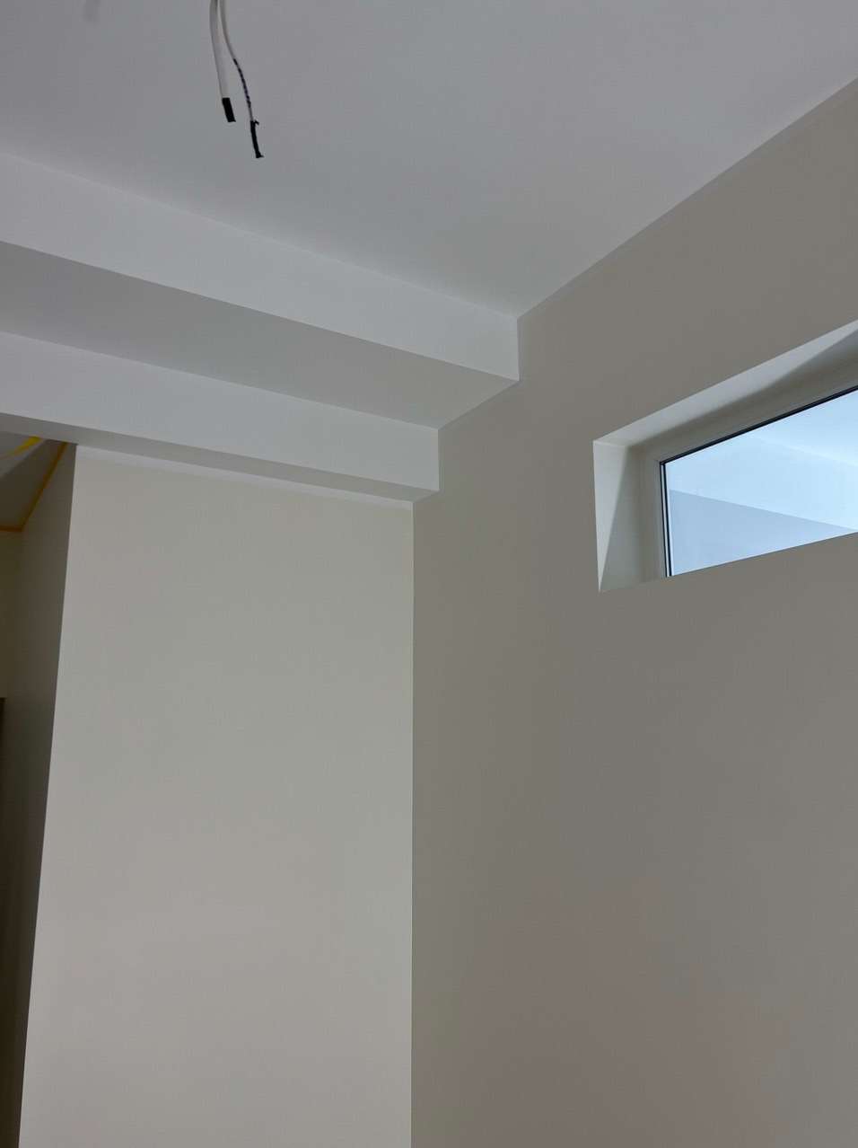 Pomieszczenie po świeżym malowaniu na kolor kremowy, z białymi elementami architektonicznymi i małym oknem. Widoczne przewody wystające z sufitu.