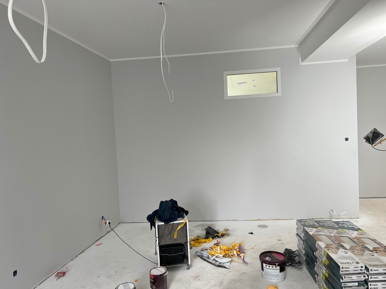 Wnętrze w trakcie odświeżania: świeżo pomalowane szare ściany, widoczne kable zwisające z sufitu, karton gips nad oknem, puszki z farbą, wózek malarski z ubraniami i paczki paneli na podłodze.