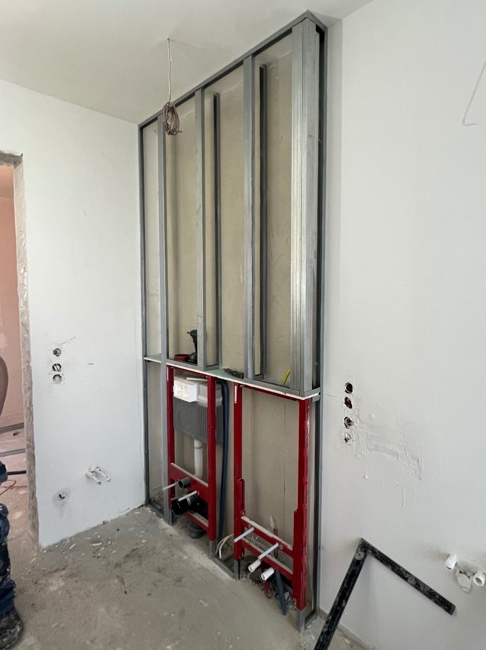 Stelaż podtynkowy WC zamontowany w metalowej konstrukcji z płyt karton gips, widoczne rury instalacyjne i wiercone otwory w ścianie pod instalacje.