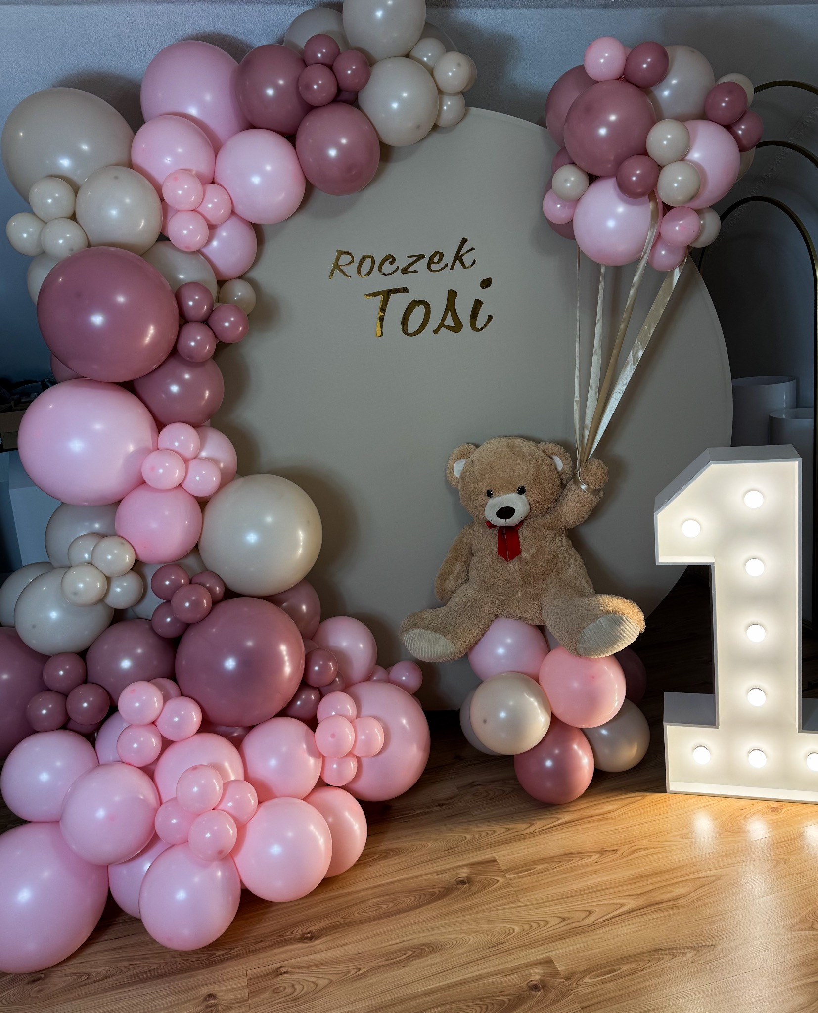 Dekoracja na roczek dziewczynki: beżowe tło z napisem 'Roczek Tosi' otoczone girlandą z różowych, białych i bordowych balonów, pluszowy miś trzymający wstążki balonów, obok podświetlana cyfra 1.