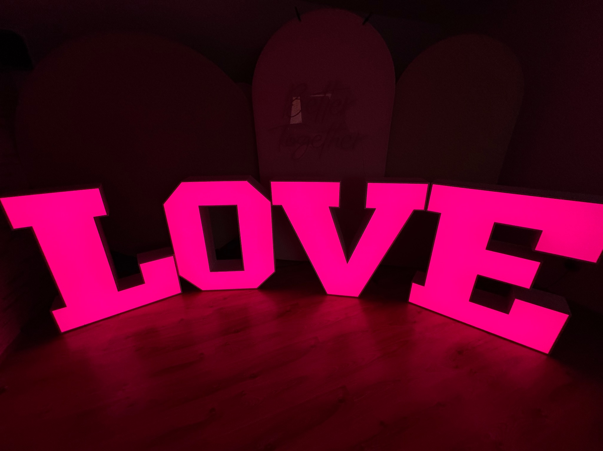 Podświetlane, neonowe litery układające się w słowo 'LOVE' w intensywnym różowym kolorze, ustawione na podłodze, z dekoracyjnym tłem. Element dekoracji weselnej lub imprezy.