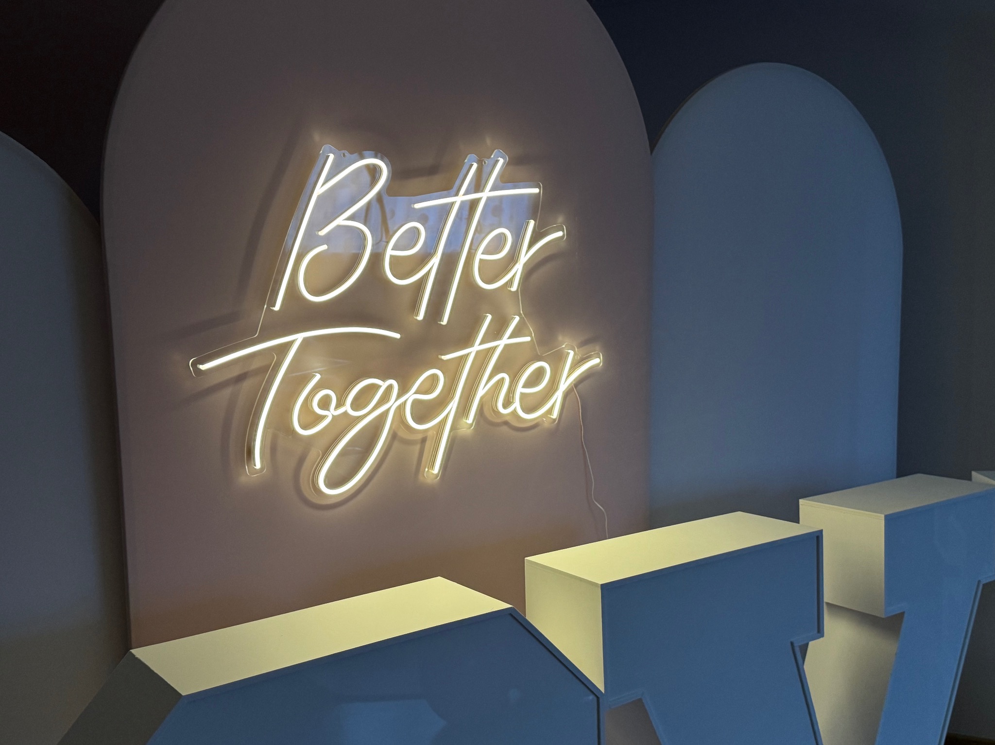 Neonowy napis 'Better Together' na tle pastelowej ściany, otoczony dekoracyjnymi elementami architektonicznymi w kształcie łuków i geometrycznych brył.