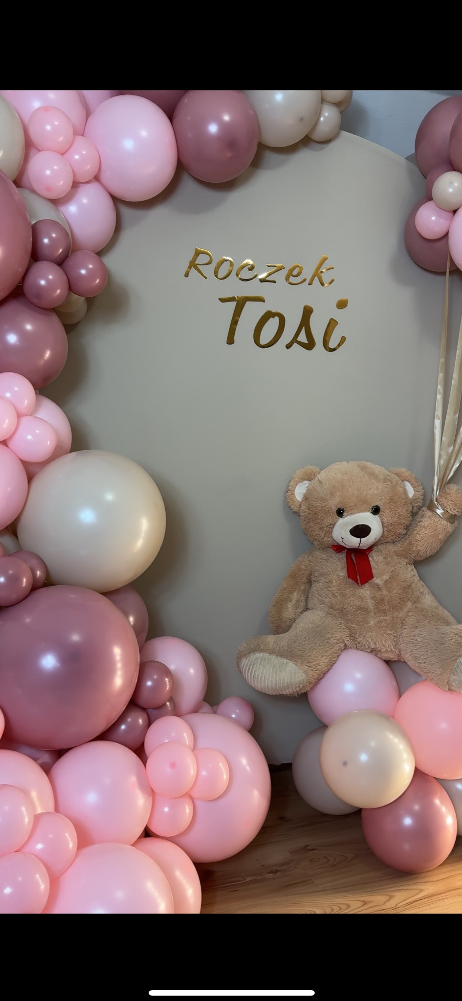 Dekoracja balonowa w odcieniach różu i beżu na roczek dziecka, z pluszowym misiem trzymającym wstążki i złotym napisem 'Roczek Tosi' na szarym tle.