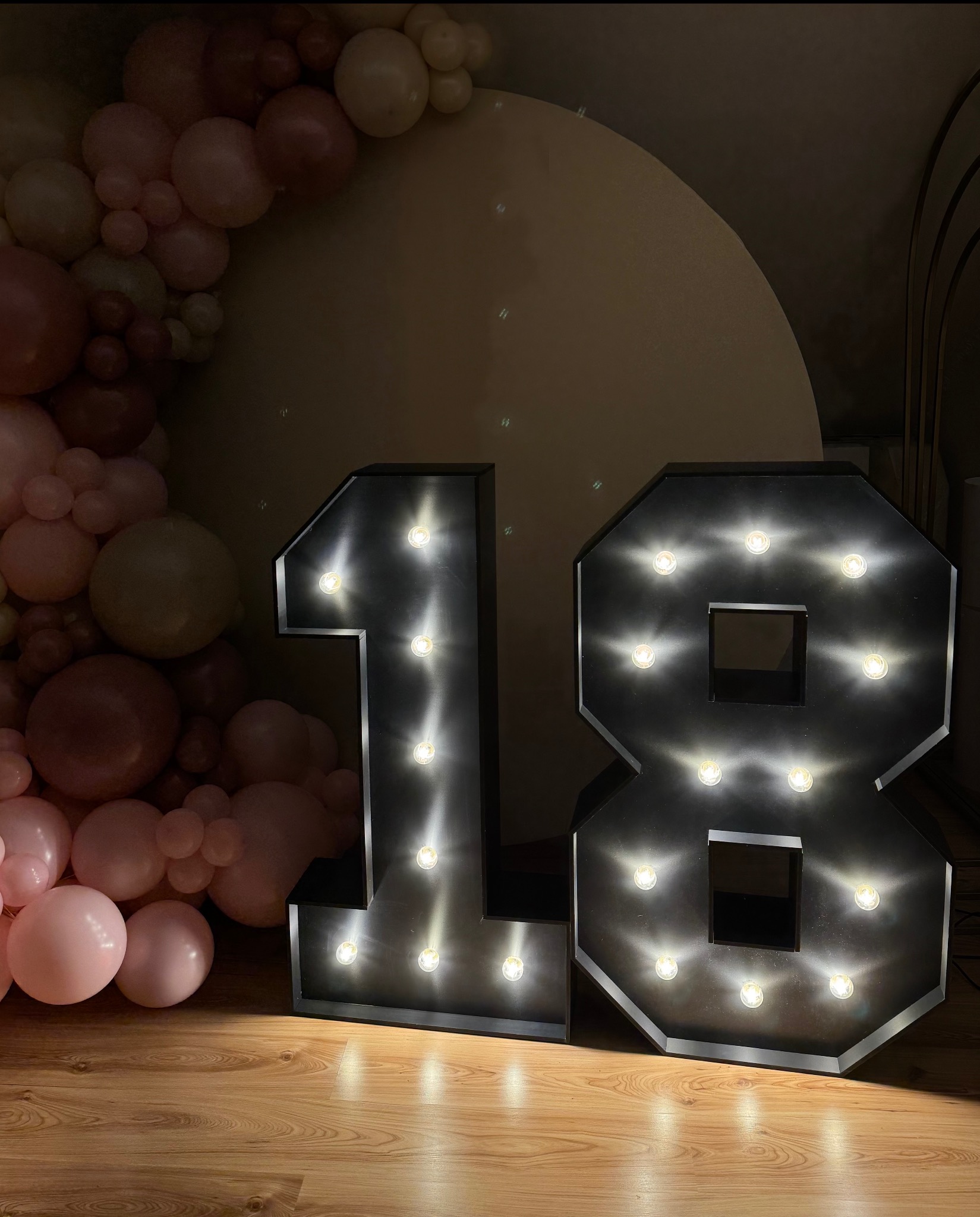 Dekoracja urodzinowa: podświetlane cyfry '18' w stylu retro z żarówkami, otoczone girlandą z różowych balonów na drewnianej podłodze.