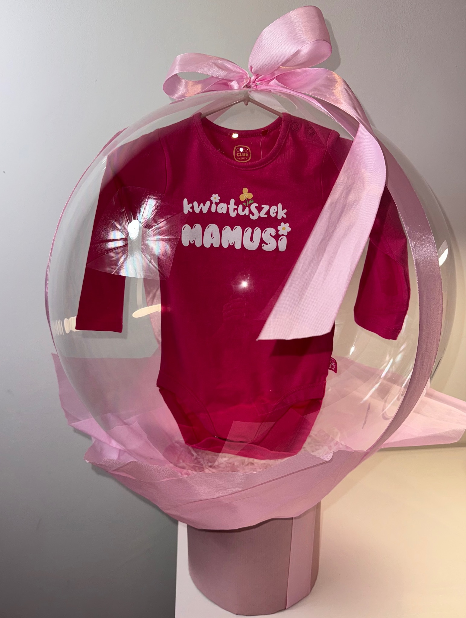 Różowe body dziecięce z napisem 'kwiatuszek mamusi' zapakowane w przezroczysty balon z różową wstążką, prezent na baby shower.