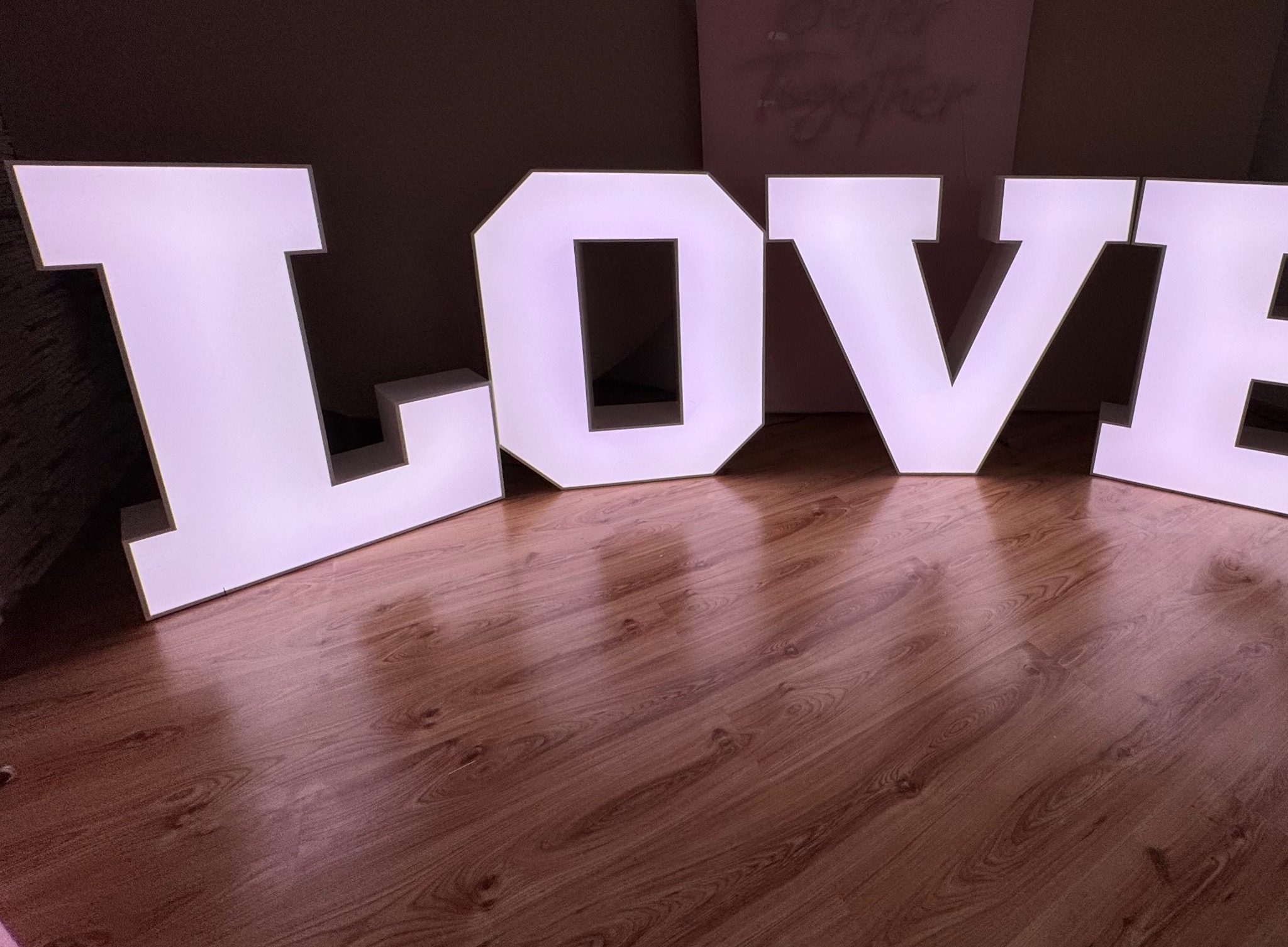 Podświetlane, białe litery LOVE na drewnianej podłodze, ustawione jako dekoracja na imprezę, z widocznym napisem 'Better Together' w tle.