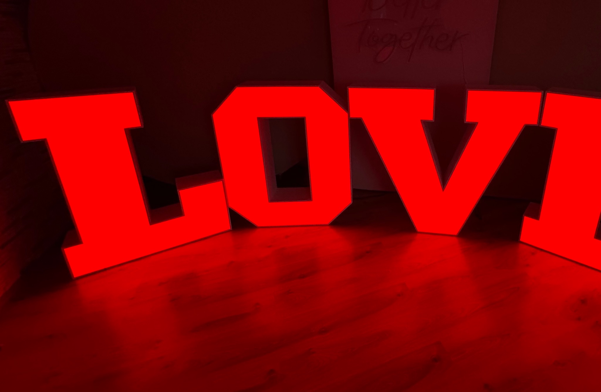 Duże, czerwone, podświetlane litery tworzące słowo 'LOVE' na błyszczącej podłodze, z delikatnym napisem 'Better Together' widocznym w tle.