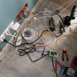 Rozłożone narzędzia i materiały do instalacji sieci komputerowej na podłodze: tester kabli, kable Ethernet różnych kolorów, wiertarka, kombinerki, zasilacz, puszka instalacyjna z wystającymi...