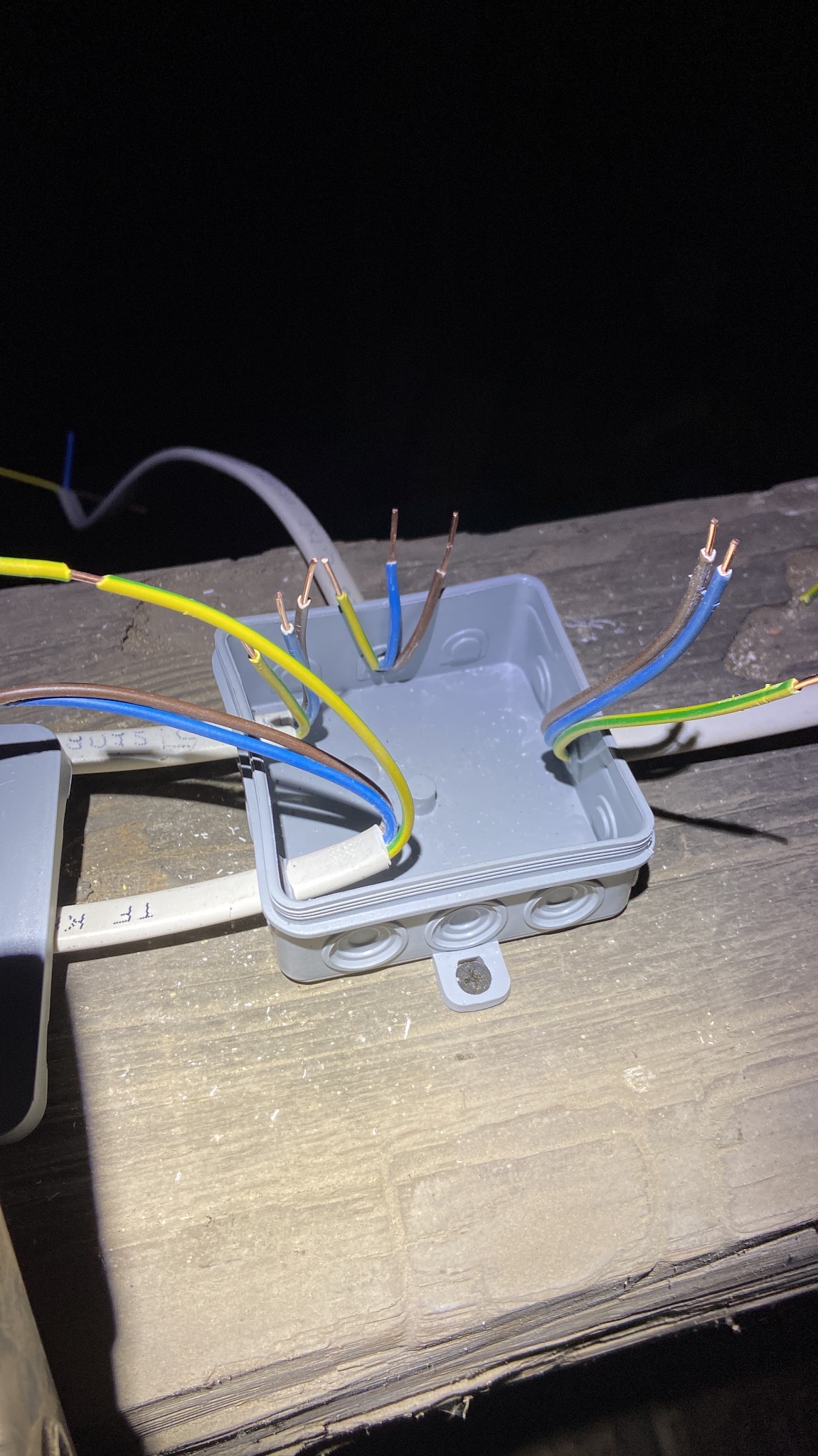 Otwarta szara puszka elektryczna z wystającymi przewodami o różnych kolorach (żółto-zielony, niebieski, brązowy) na tle surowego, drewnianego podłoża, widok z góry.