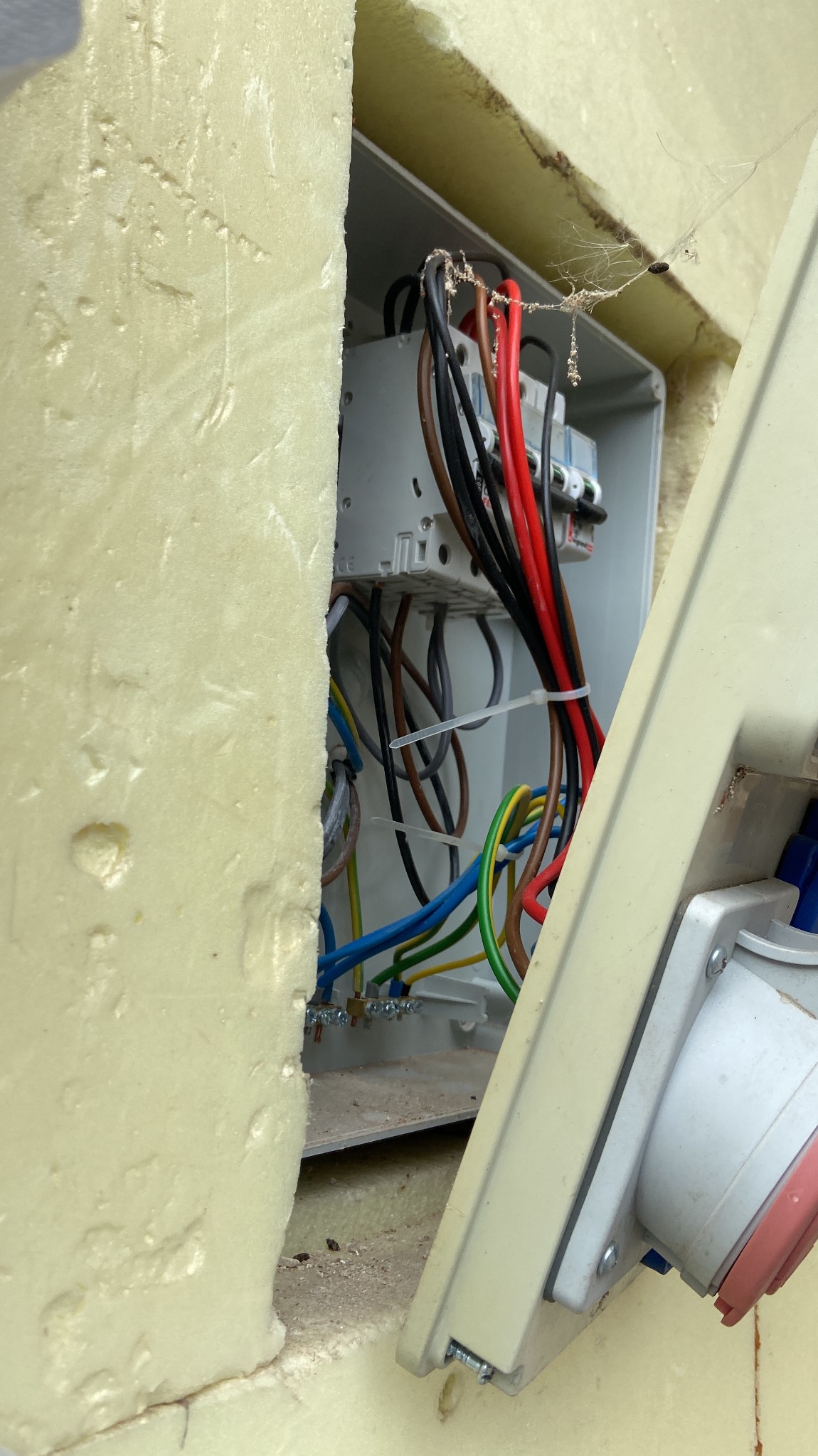 Otwarta skrzynka elektryczna z widocznymi przewodami w kolorach czerwonym, czarnym, brązowym, niebieskim i zielono-żółtym, zabezpieczonymi opaskami zaciskowymi, umieszczona w ścianie ocieplonej...