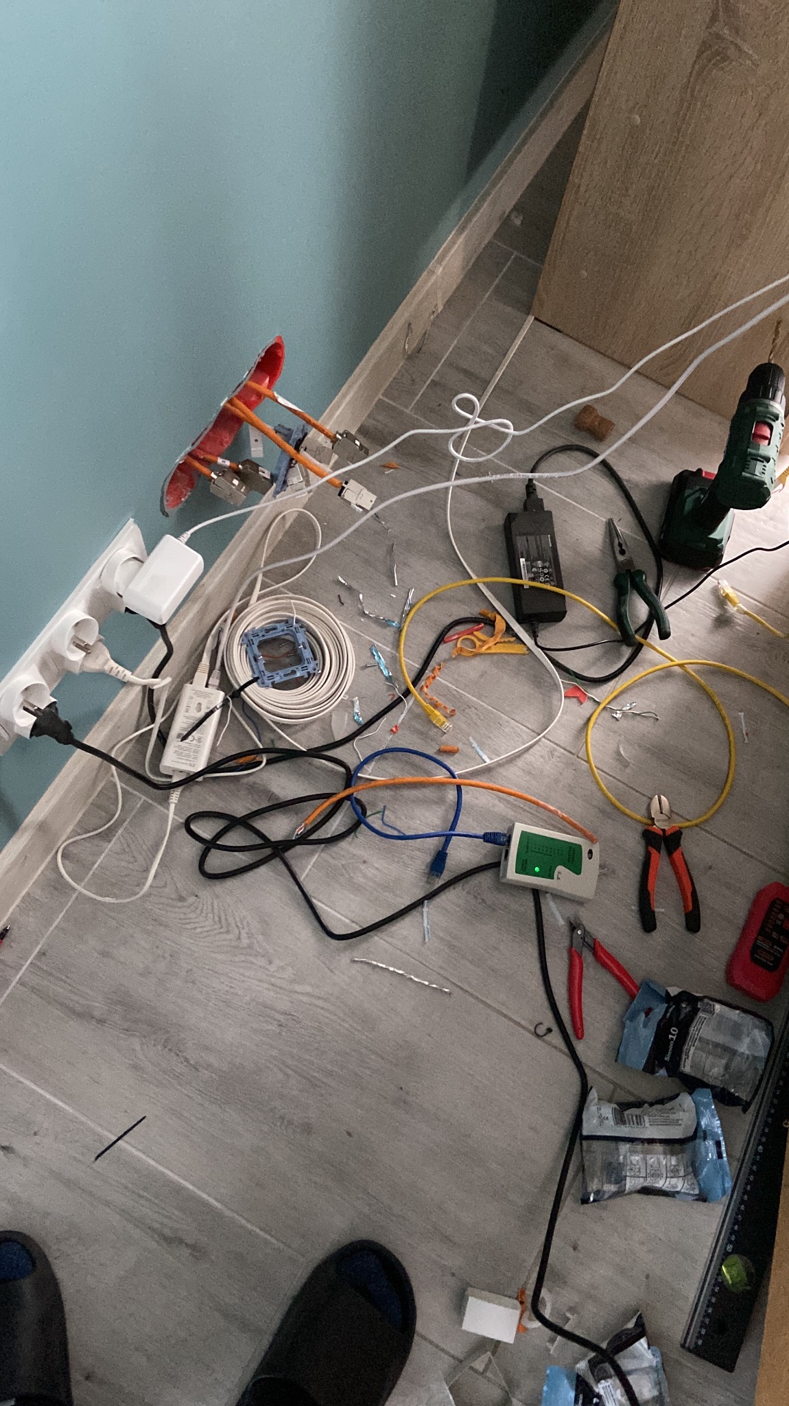 Rozłożone narzędzia i materiały do instalacji sieci komputerowej na podłodze: tester kabli, kable Ethernet różnych kolorów, wiertarka, kombinerki, zasilacz, puszka instalacyjna z wystającymi...