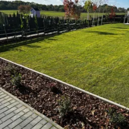 Zadbany ogród z równo przystrzyżonym trawnikiem, obrzeżem z kory i nasadzeniami, w tle zielony krajobraz z domkiem ogrodowym i drzewami o jesiennych barwach.