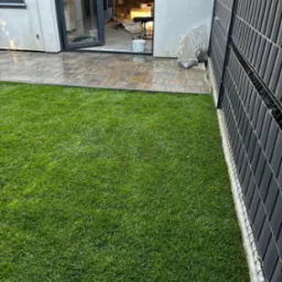 Świeżo założony trawnik z rolki przylegający do kostki brukowej i nowoczesnego ogrodzenia panelowego. W tle widoczne otwarte drzwi tarasowe i wnętrze domu w trakcie wykończenia.