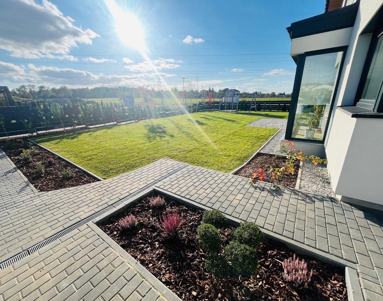 Widok na zadbany ogród z kostką brukową, rabatami z wrzosami i kulistymi krzewami, oraz trawnikiem z placem zabaw w tle, oświetlony promieniami słonecznymi.