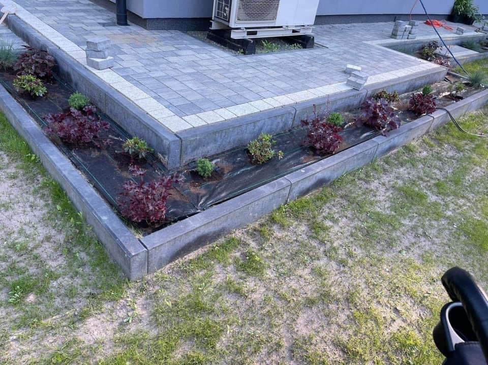 Kaskadowy taras z kostki brukowej z obrzeżami, rabata z agrowłókniną i młodymi roślinami, widok z góry na fragment trawnika.