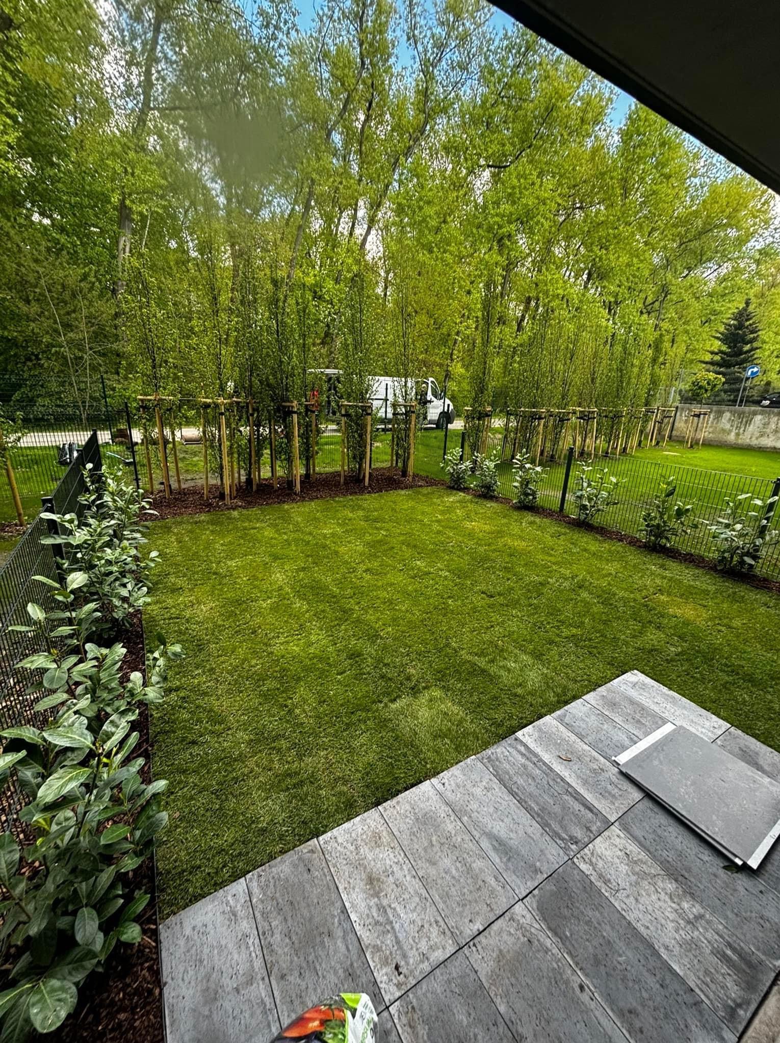 Zadbany trawnik z nowo posadzonymi drzewami, ogrodzony siatką, z widocznym fragmentem tarasu z płyt kamiennych i samochodem dostawczym w tle.