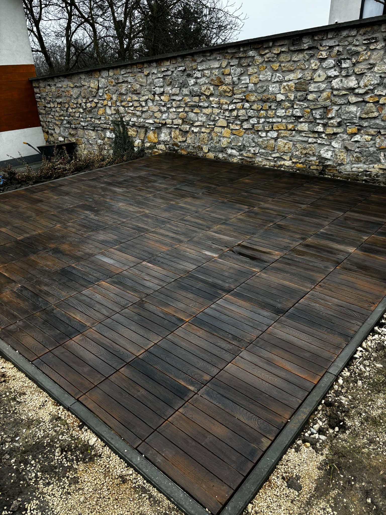 Ułożona nawierzchnia z ciemnobrązowych, kwadratowych płyt imitujących drewno, obrzeże z szarego betonu, podsypka z jasnego żwiru, w tle kamienny mur.