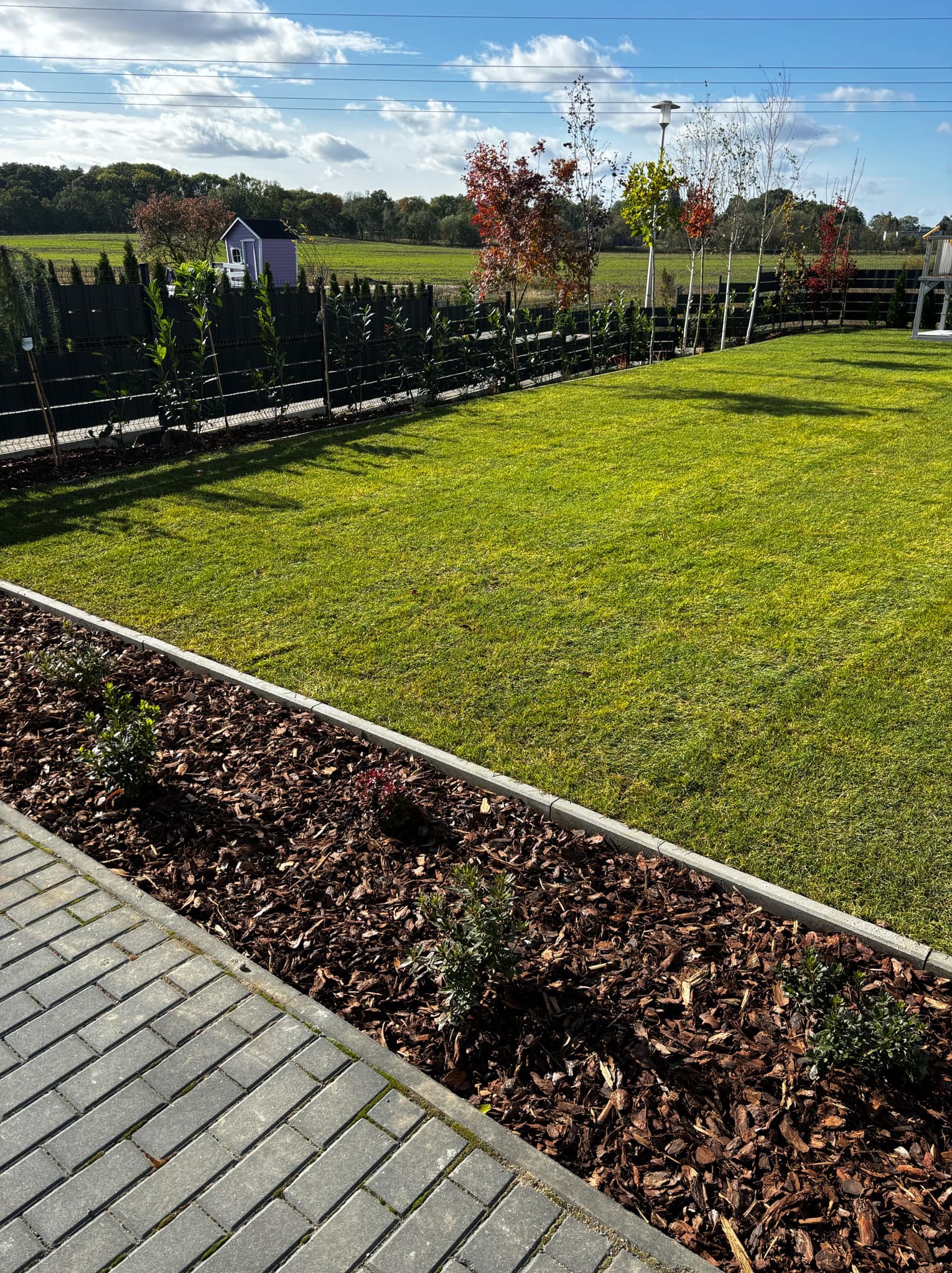 Zadbany ogród z równo przystrzyżonym trawnikiem, obrzeżem z kory i nasadzeniami, w tle zielony krajobraz z domkiem ogrodowym i drzewami o jesiennych barwach.