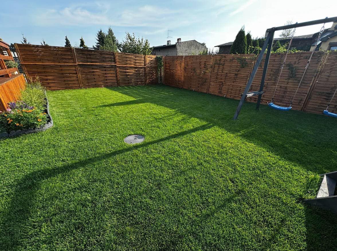 Świeżo założony trawnik z rolki, ogrodzony drewnianym płotem, z widocznym placem zabaw z huśtawkami i okrągłą studzienką kanalizacyjną pośrodku.