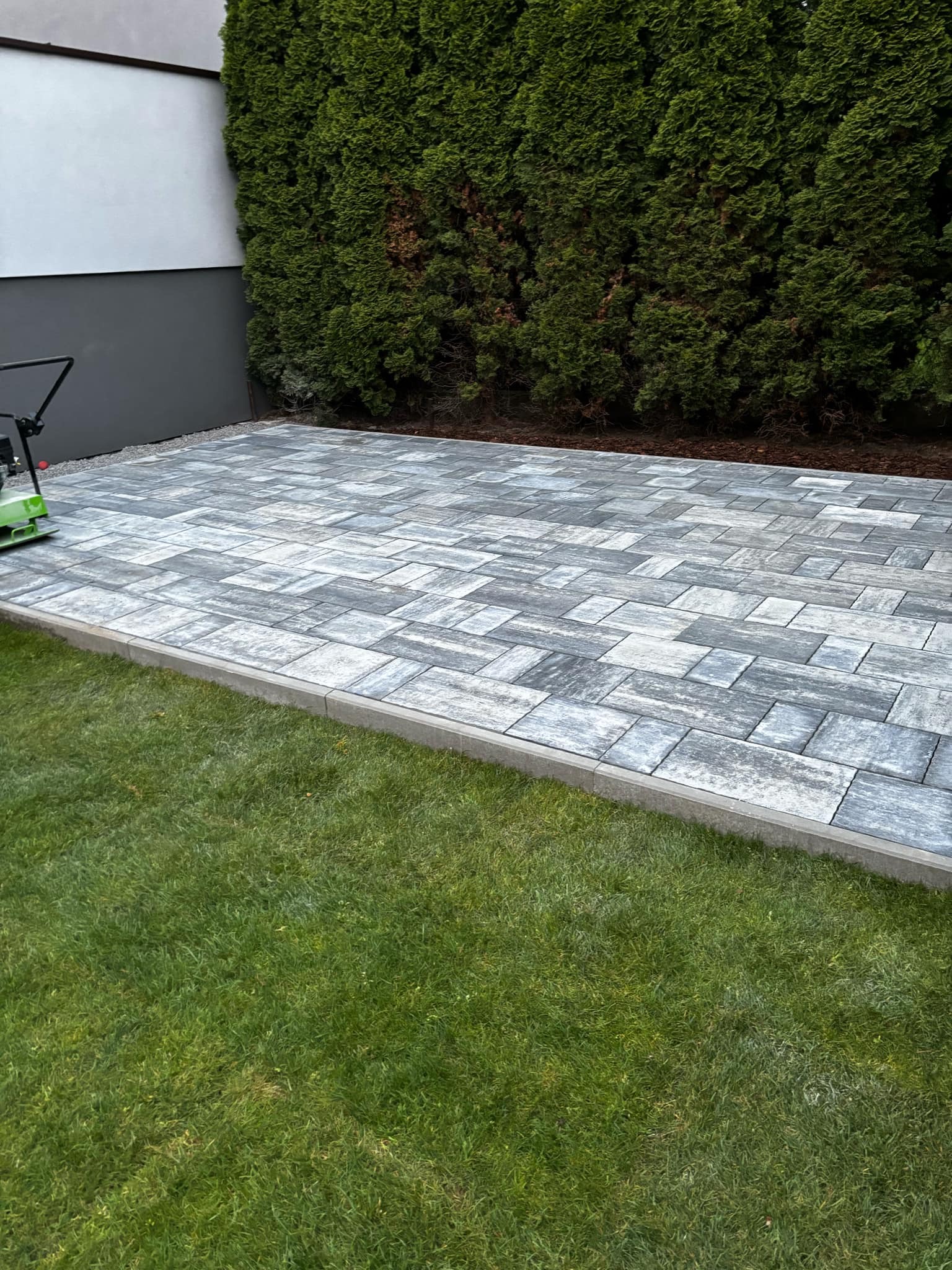 Świeżo ułożony chodnik z szarej kostki brukowej o nieregularnym wzorze, obrzeża z betonowych krawężników, obok trawnik i wysokie tuje, w tle ściana budynku. Widoczna zagęszczarka płytowa.