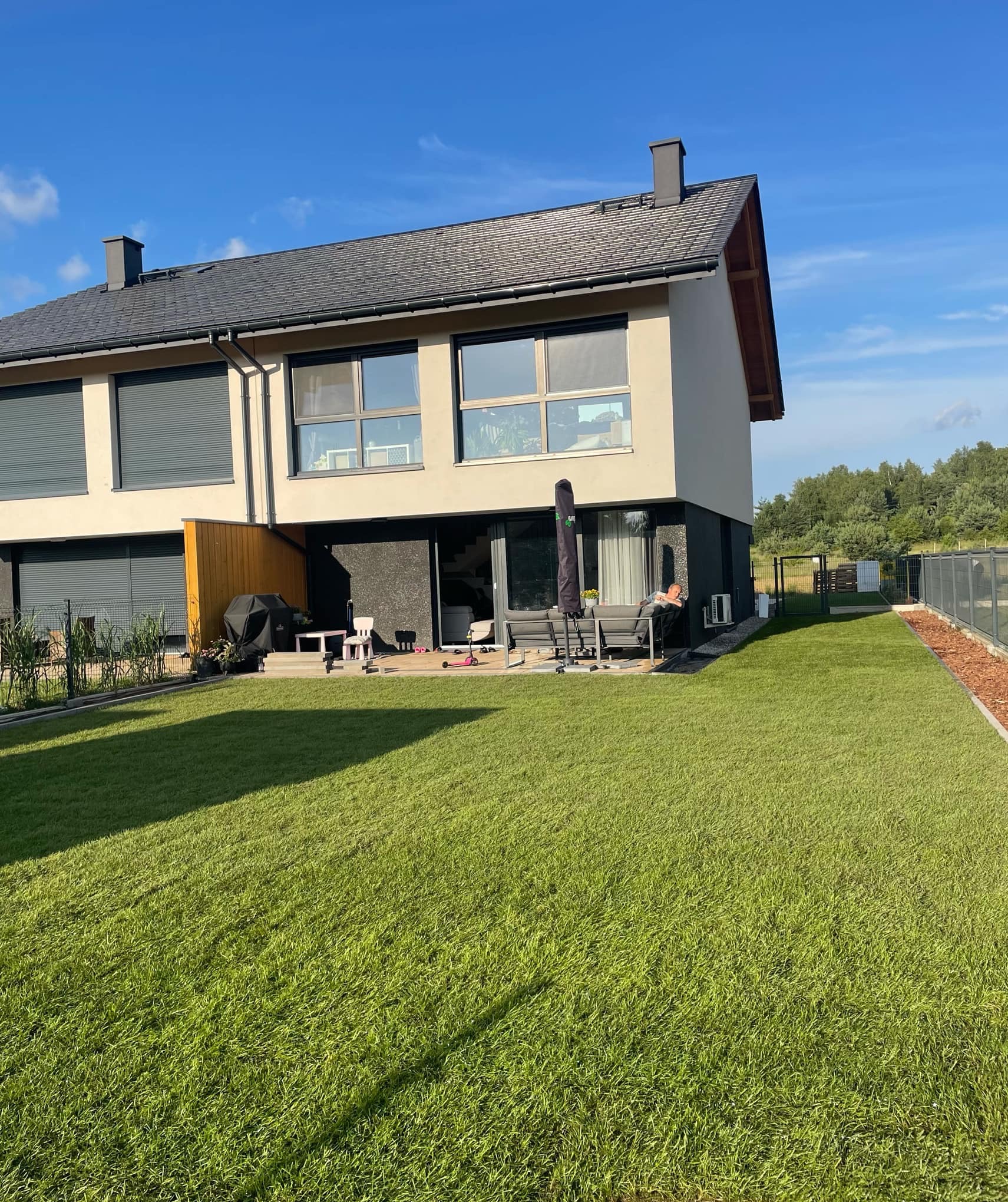 Widok na dom z zadbanym trawnikiem z rolki, tarasem i meblami ogrodowymi w słoneczny dzień.