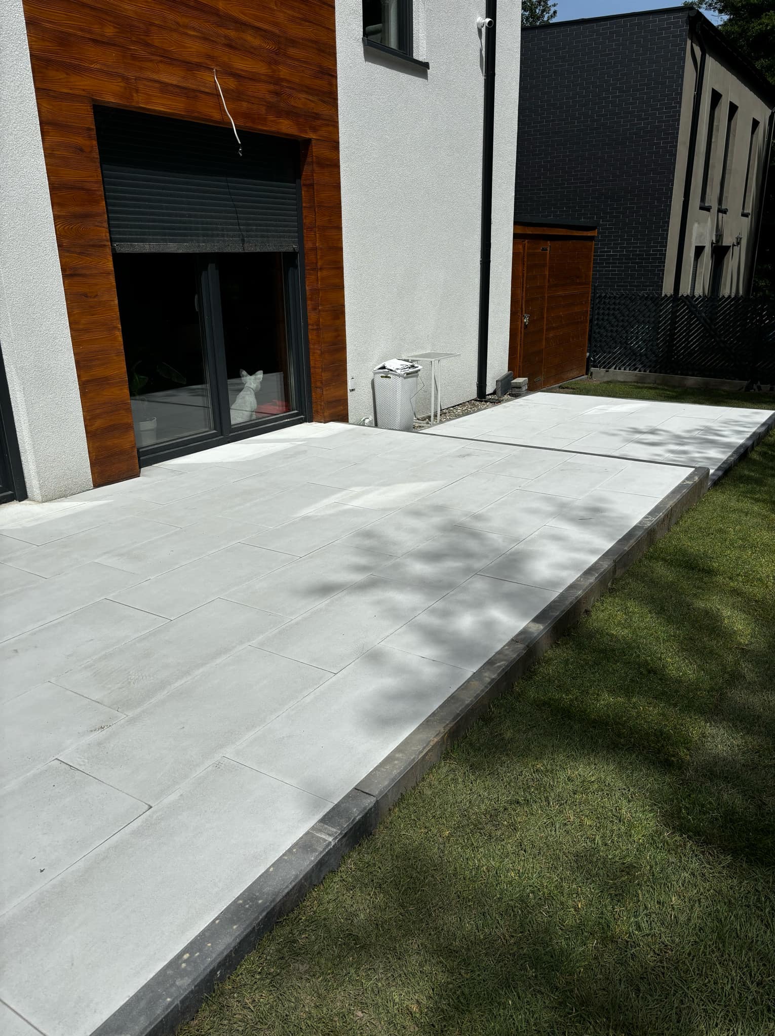 Nowoczesny taras z jasnoszarej kostki brukowej, obrzeża z ciemnoszarego kamienia, widok na trawnik i budynek z drewnianą elewacją, w oknie siedzi biały kot.