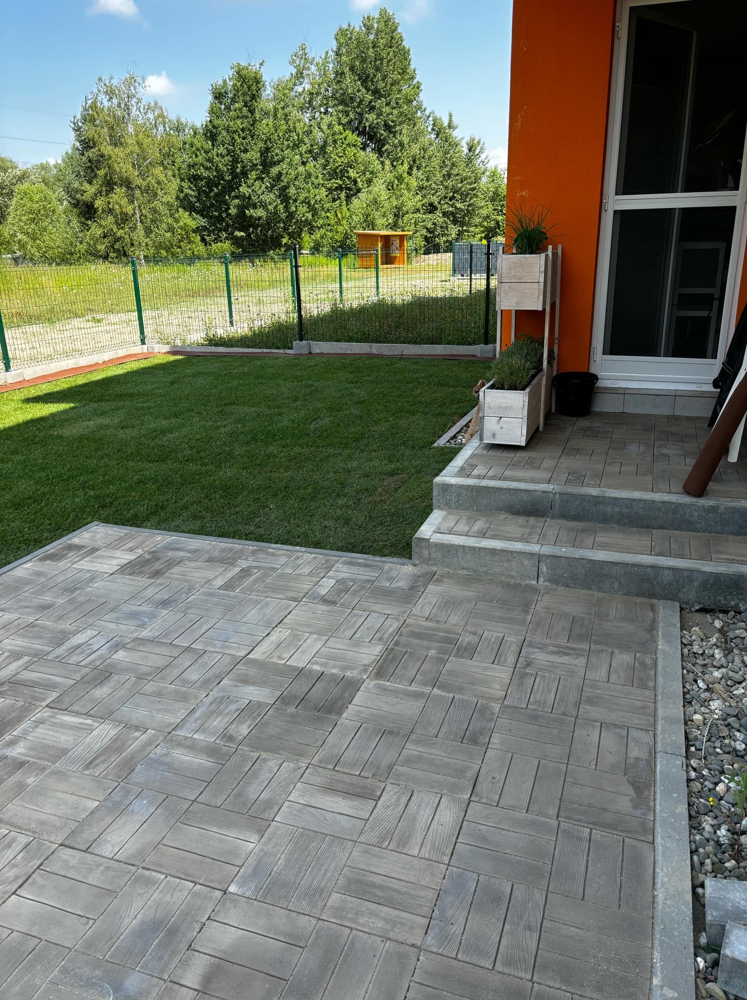 Nowo ułożona, szara kostka brukowa z wzorem drewna na podjeździe, obok świeżo założony trawnik i pomarańczowy budynek z białymi ramami okiennymi.