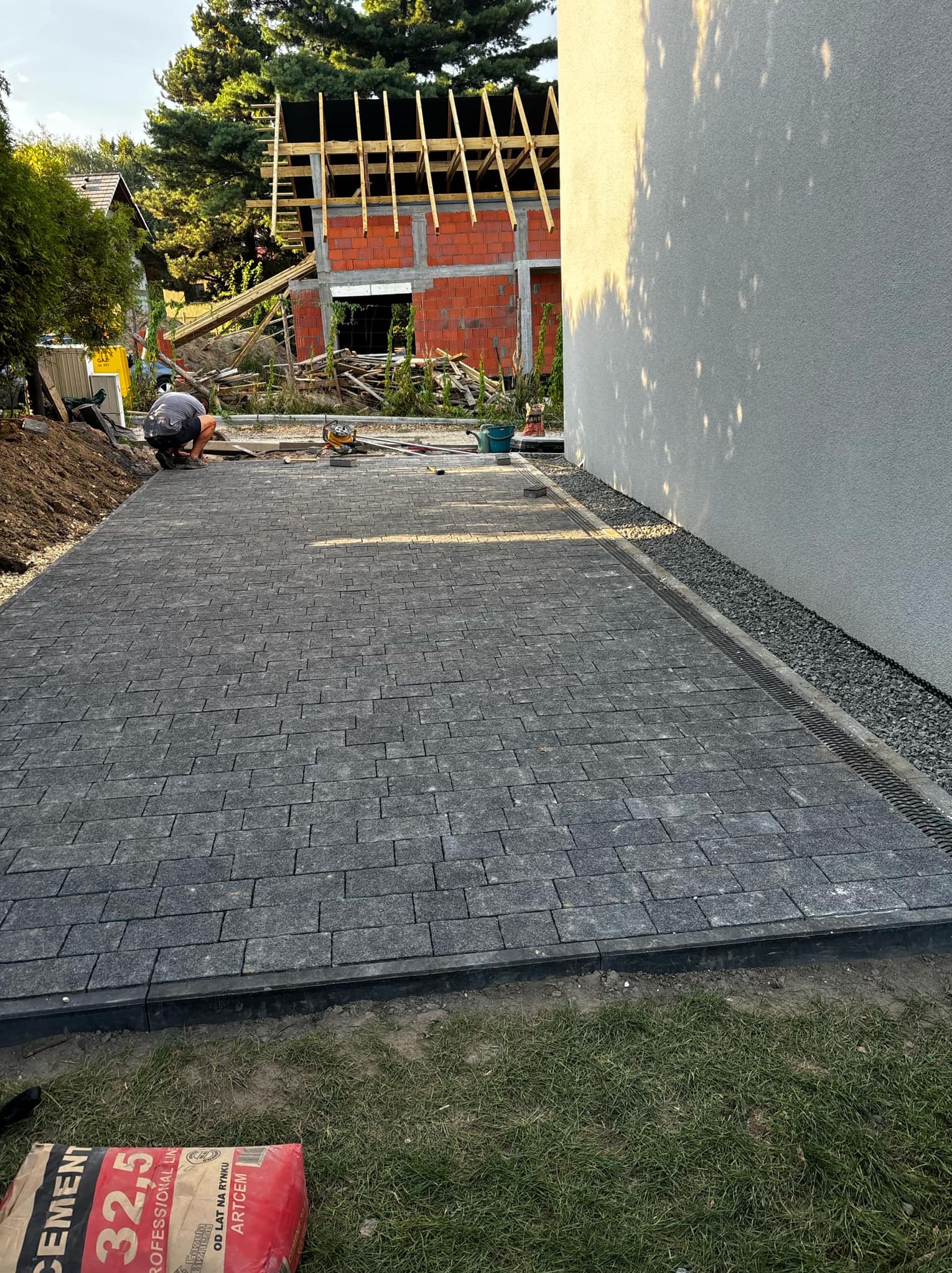 Układanie szarej kostki brukowej na posesji, widoczny pracownik w trakcie pracy, obok ściana budynku i worki z cementem.