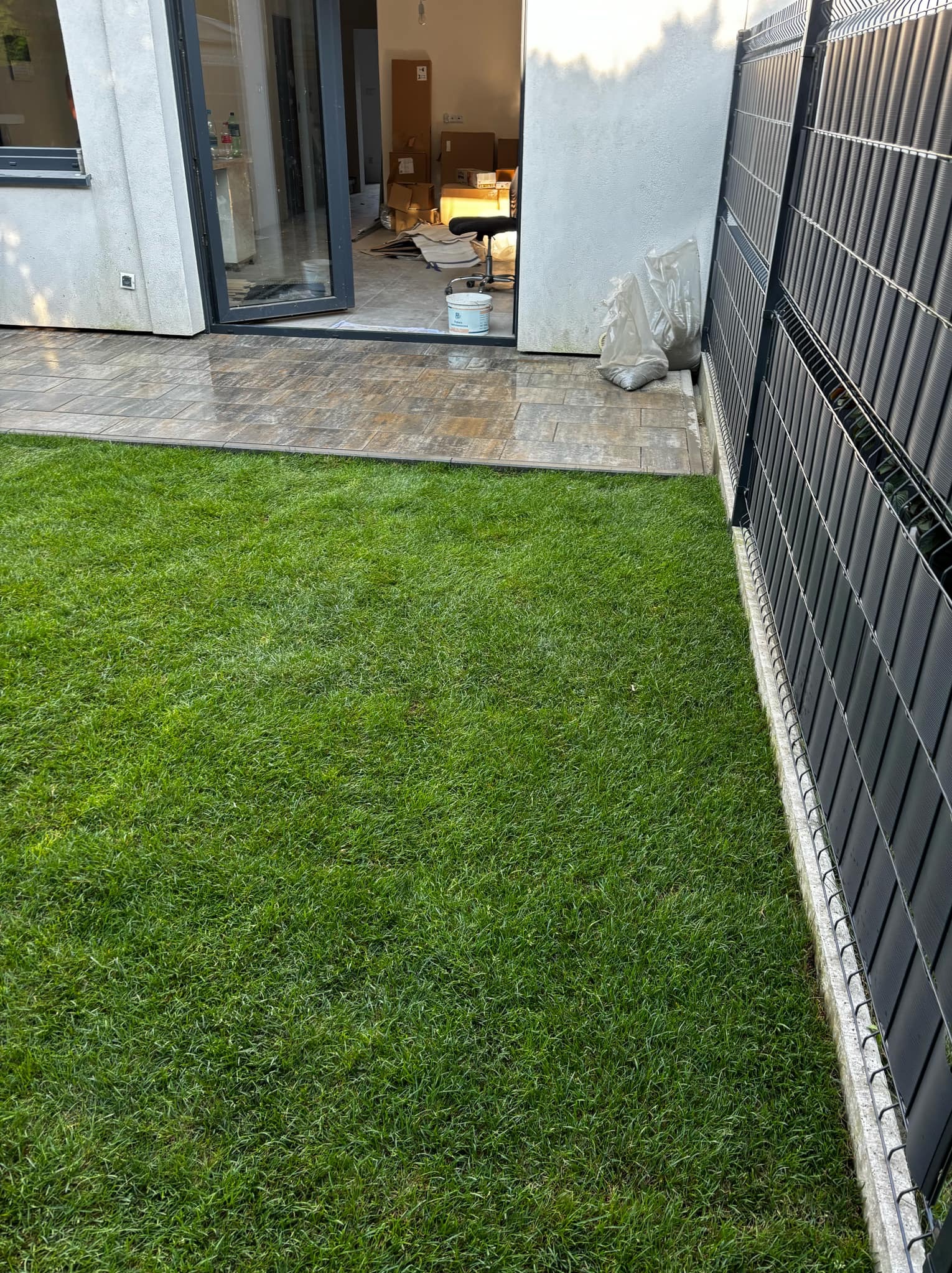 Świeżo założony trawnik z rolki przylegający do kostki brukowej i nowoczesnego ogrodzenia panelowego. W tle widoczne otwarte drzwi tarasowe i wnętrze domu w trakcie wykończenia.
