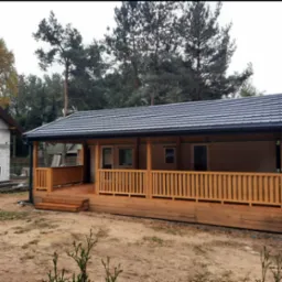 Nowoczesny dom z drewnianą werandą i grafitowym dachem z blachodachówki modułowej, otoczony drzewami.