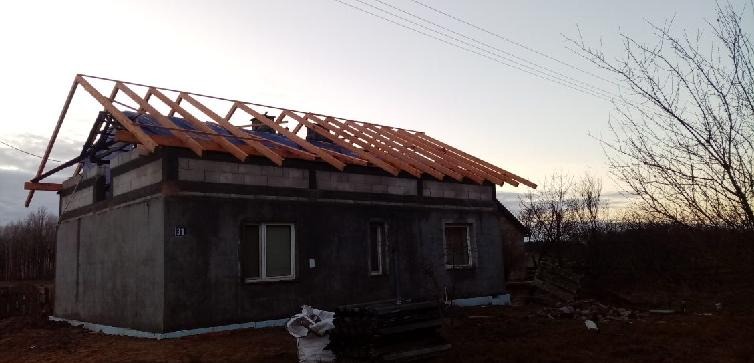 Budynek w trakcie budowy z widoczną konstrukcją dachu z drewnianych krokwi i częściowo położoną membraną dachową, w tle zachodzące słońce.