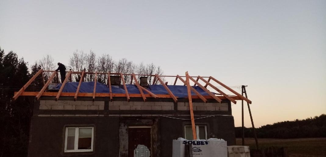 Montaż drewnianej konstrukcji dachu na budynku z pustaków, zabezpieczonej niebieską plandeką, pracownik na dachu, w tle drzewa i zachmurzone niebo.