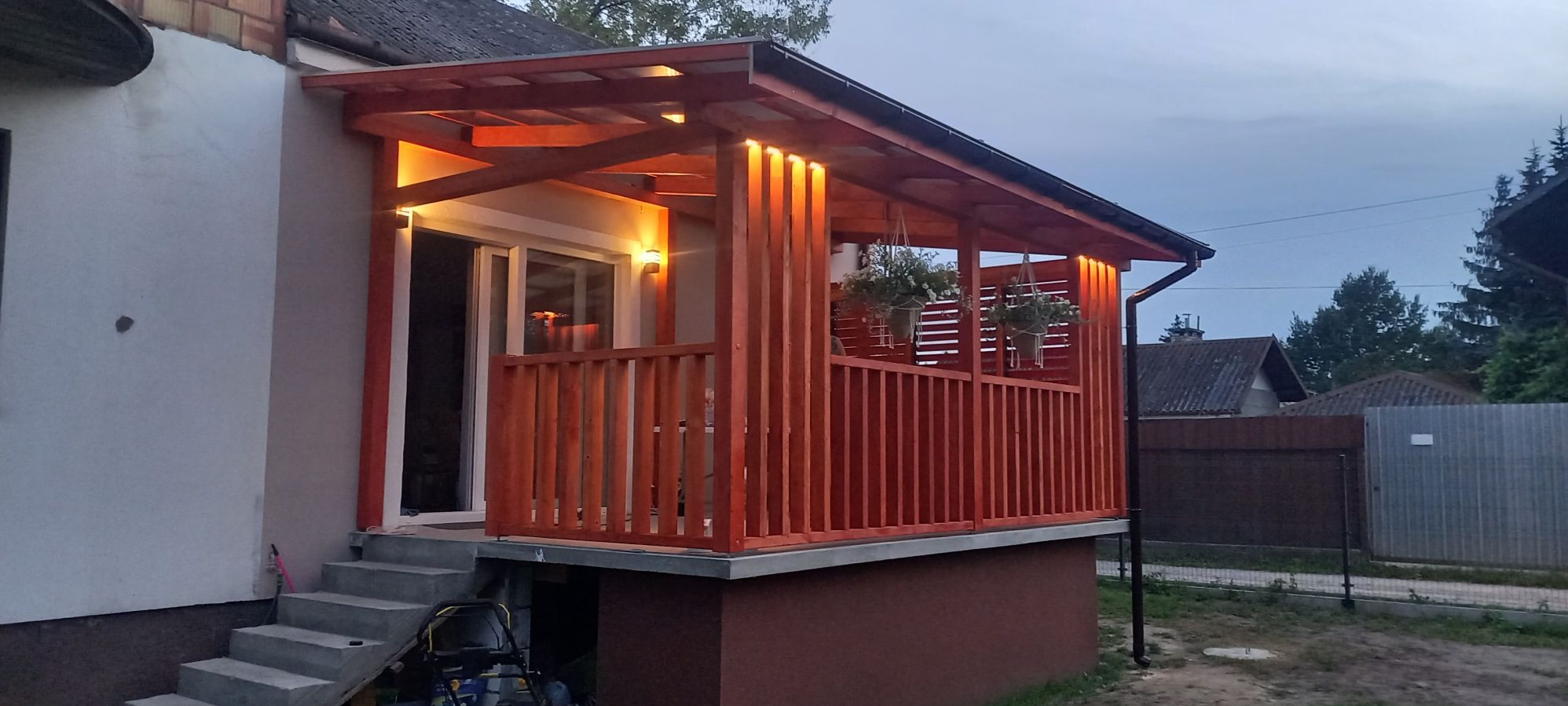 Zadaszony taras z drewnianą balustradą, oświetleniem LED i wiszącymi donicami z kwiatami, przylegający do domu ze schodami wejściowymi.