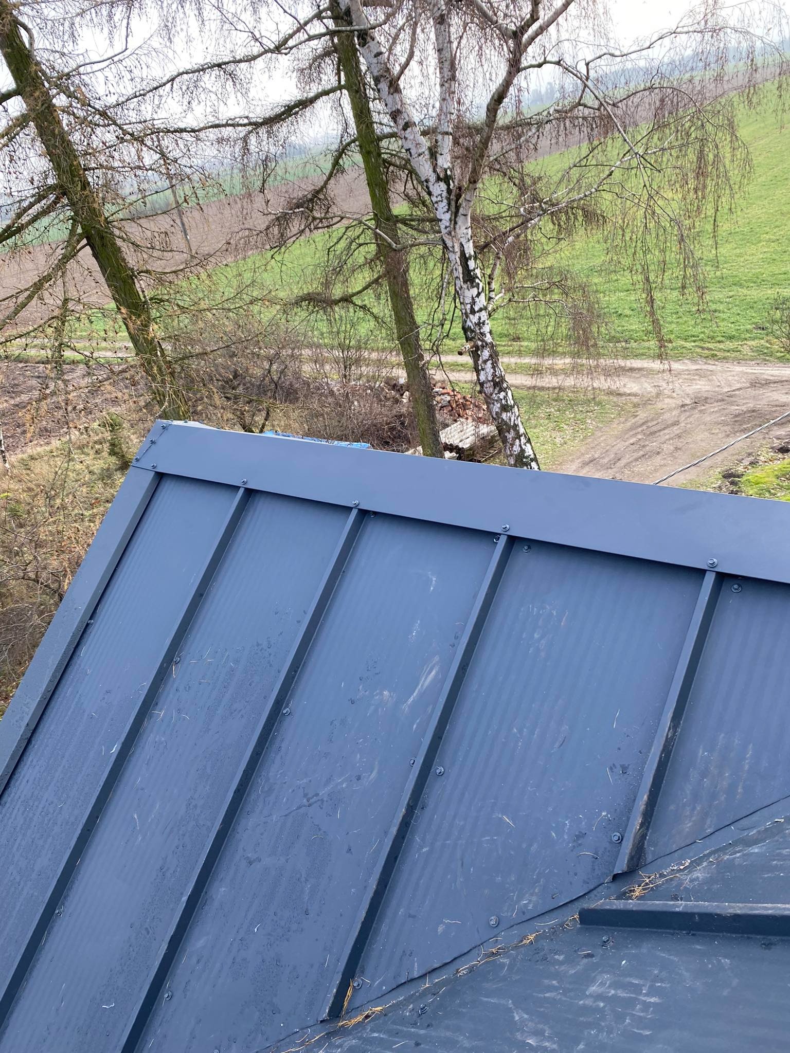 Nowy, grafitowy dach z blachy na rąbek stojący, widok z góry, z widocznymi drzewami i polami w tle.