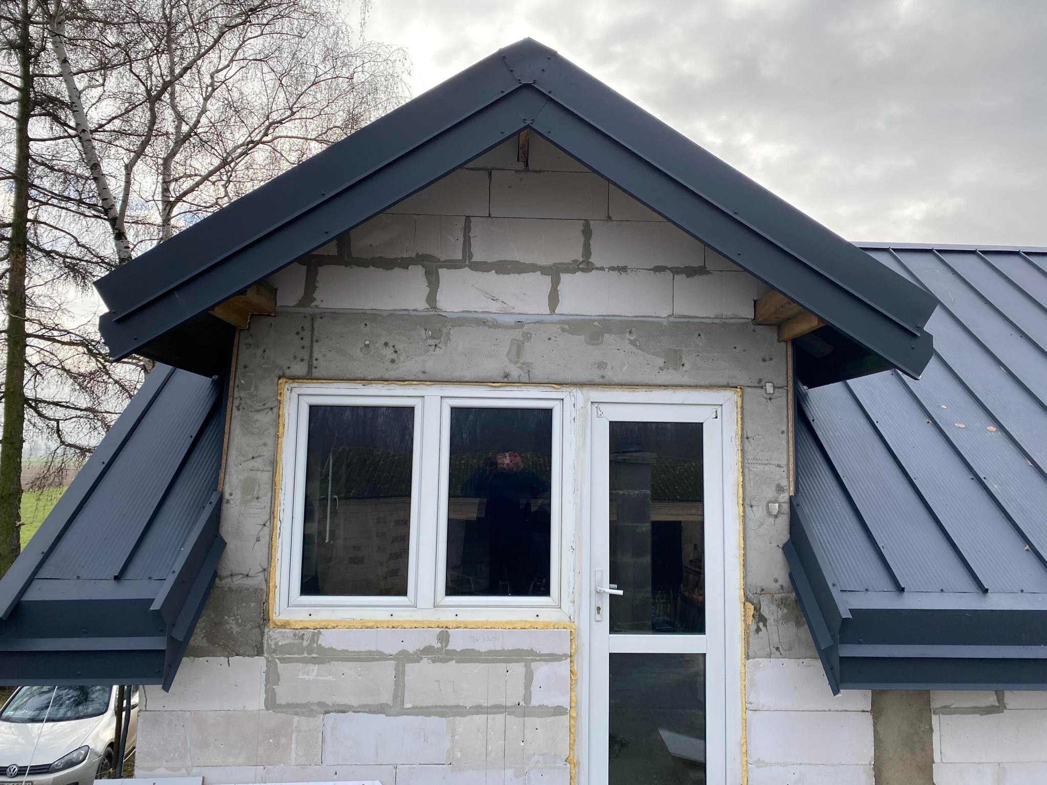 Fragment domu w trakcie budowy z zamontowaną, grafitową blachodachówką na dachu, widoczne okno i drzwi balkonowe, ściany z betonu komórkowego.