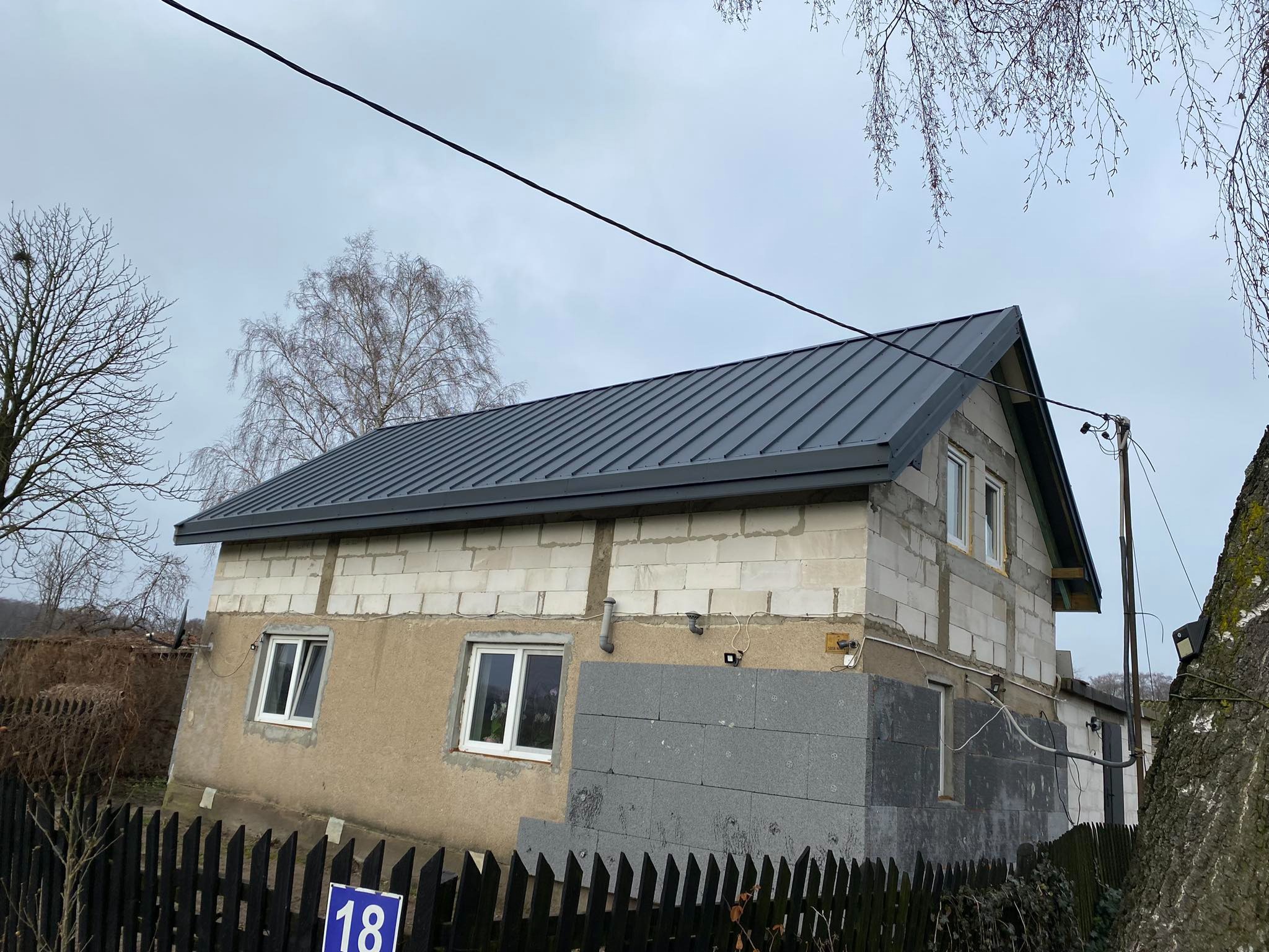 Dom w trakcie budowy z nowym, grafitowym dachem z blachy trapezowej, widoczne białe bloczki i szara elewacja, drewniane ogrodzenie w tle.