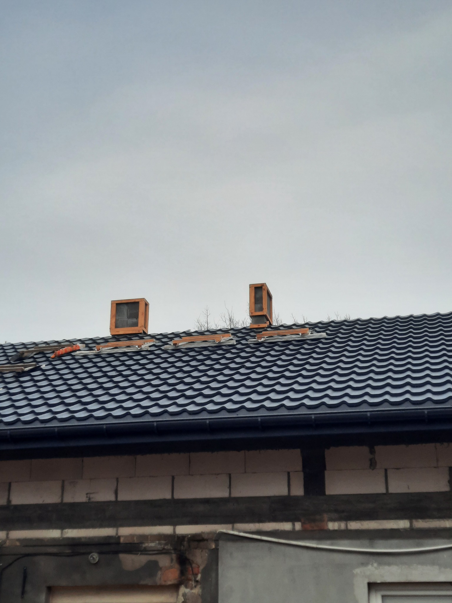 Nowy, czarny dach z blachodachówki z dwoma kominami w trakcie montażu, widoczne elementy rusztowania komina i fragmenty starego pokrycia dachowego, szare niebo w tle.