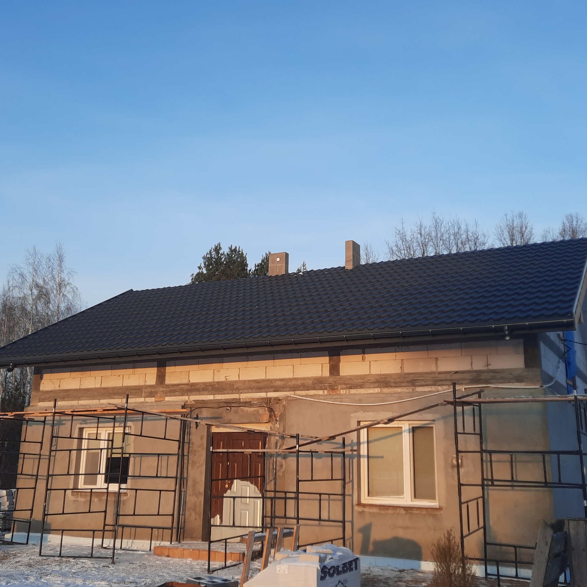 Dom w trakcie remontu z nowym, czarnym dachem z blachodachówki, widoczne rusztowania i materiały budowlane na posesji.