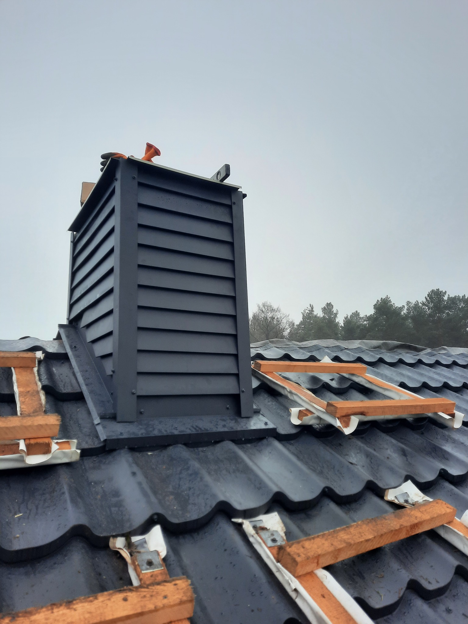 Nowy, grafitowy komin wentylacyjny z drewnianą obudową, zamontowany na dachu z czarnej dachówki ceramicznej. Widoczne drewniane elementy mocujące dachówki. W tle zalesione wzgórze pod pochmurnym...