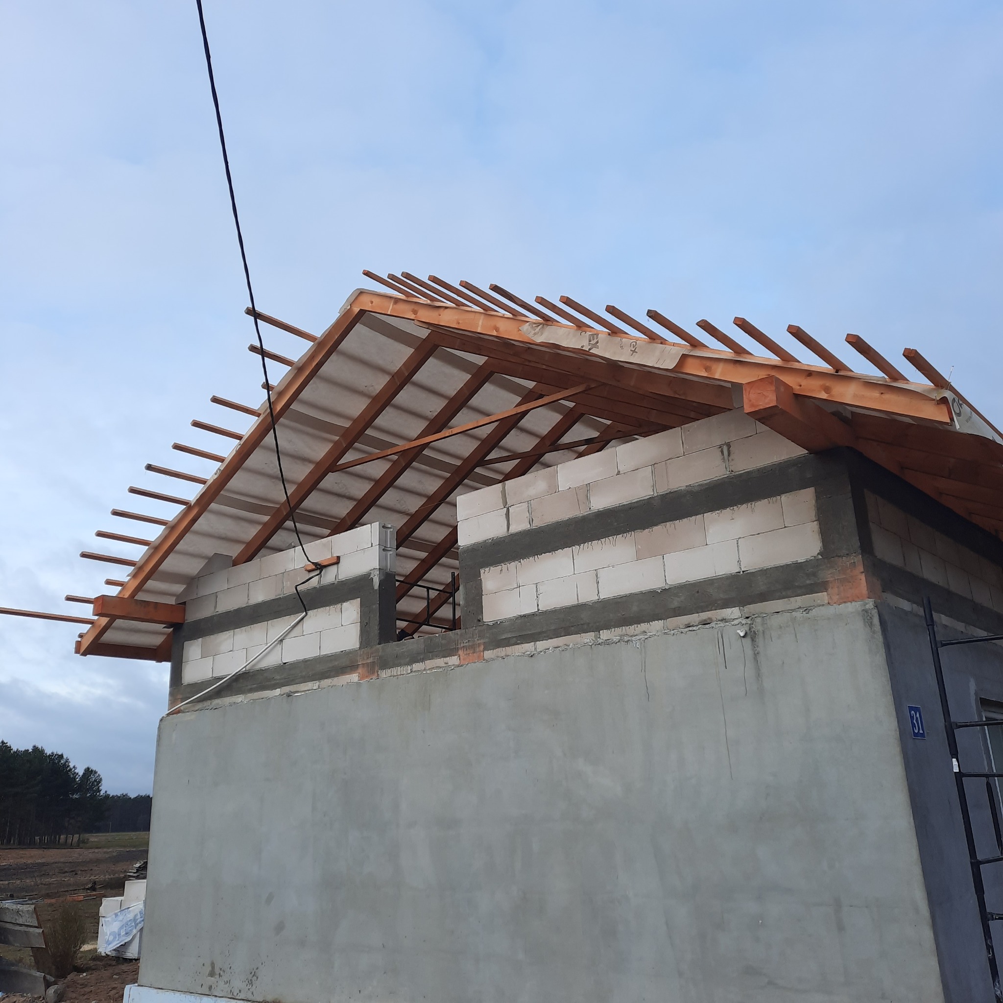 Wznoszenie konstrukcji drewnianej dachu na budynku z betonu komórkowego, widoczne krokwie i membrana dachowa, na tle pochmurnego nieba.