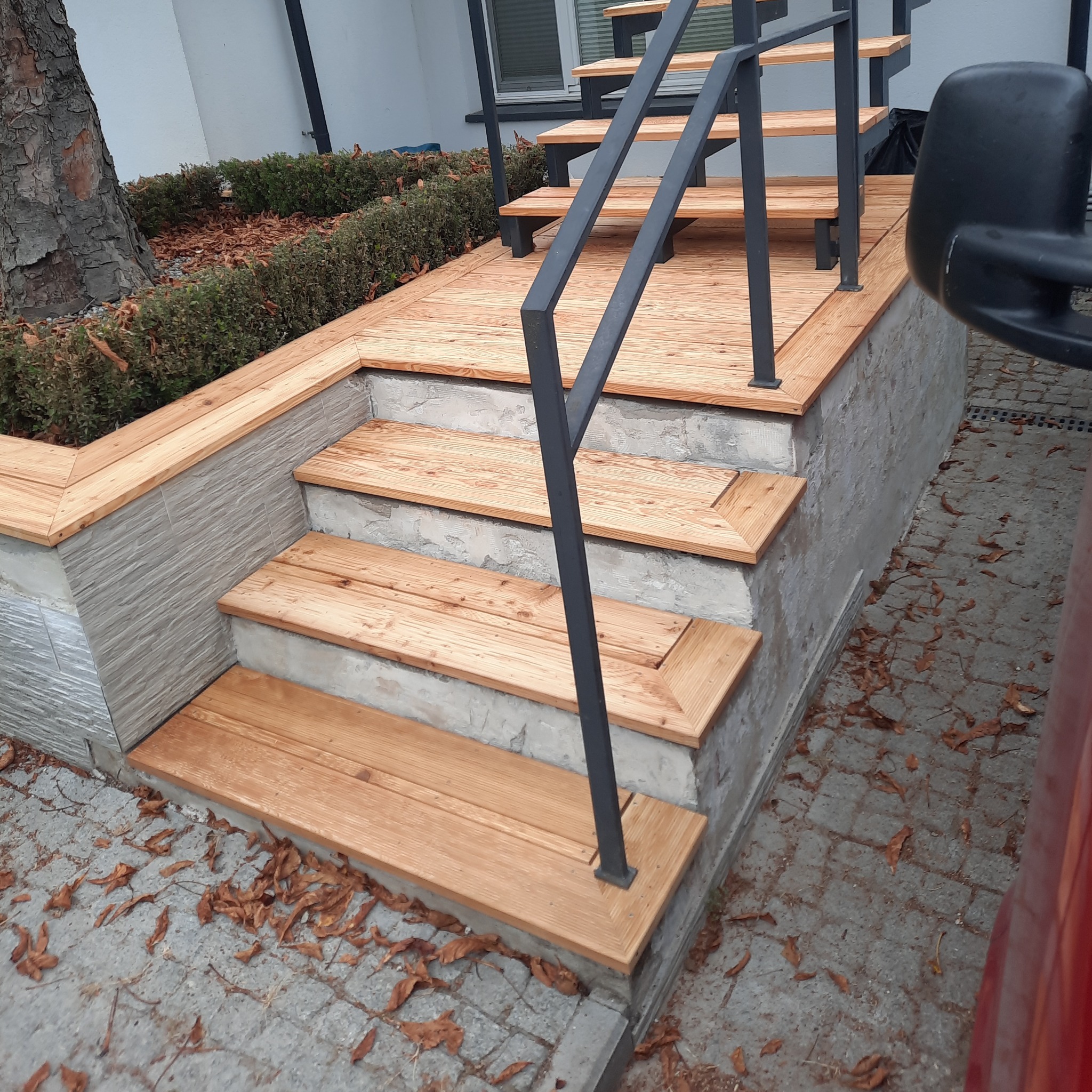 Zewnętrzne schody z drewna z szarą balustradą, wykończone płytkami imitującymi kamień, prowadzące do podwyższonego tarasu, otoczone zielenią i opadłymi liśćmi.