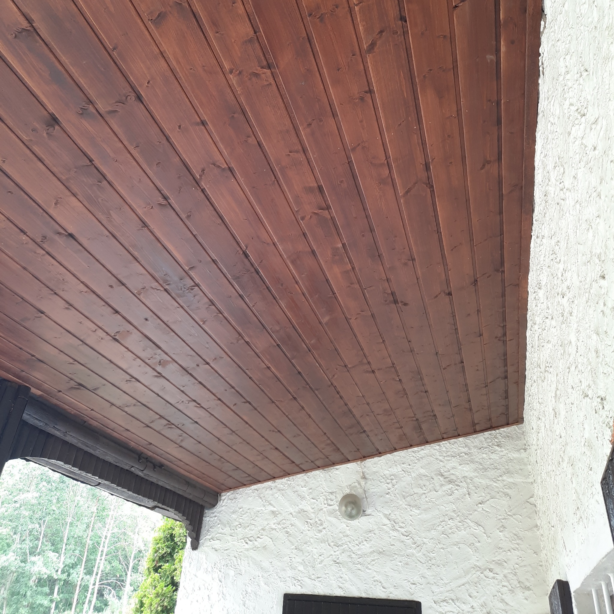 Zadaszenie tarasu wykończone ciemnobrązową boazerią drewnianą, widok od dołu, biały tynk strukturalny ściany.